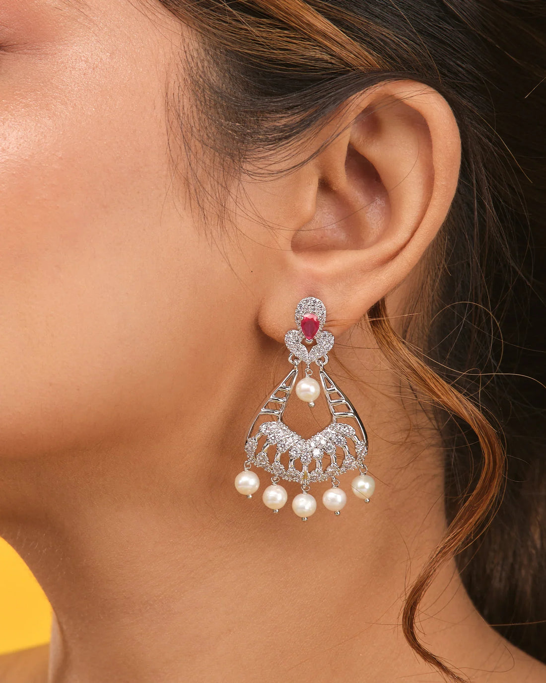 The Regal Pearl Cascade Jhumka by Chandrani Pearls features silver CZ work, intricate designs, pink stones at the top, and pearl drops—ideal wedding jewelry—shown on a tan surface with orange fabric in the background.