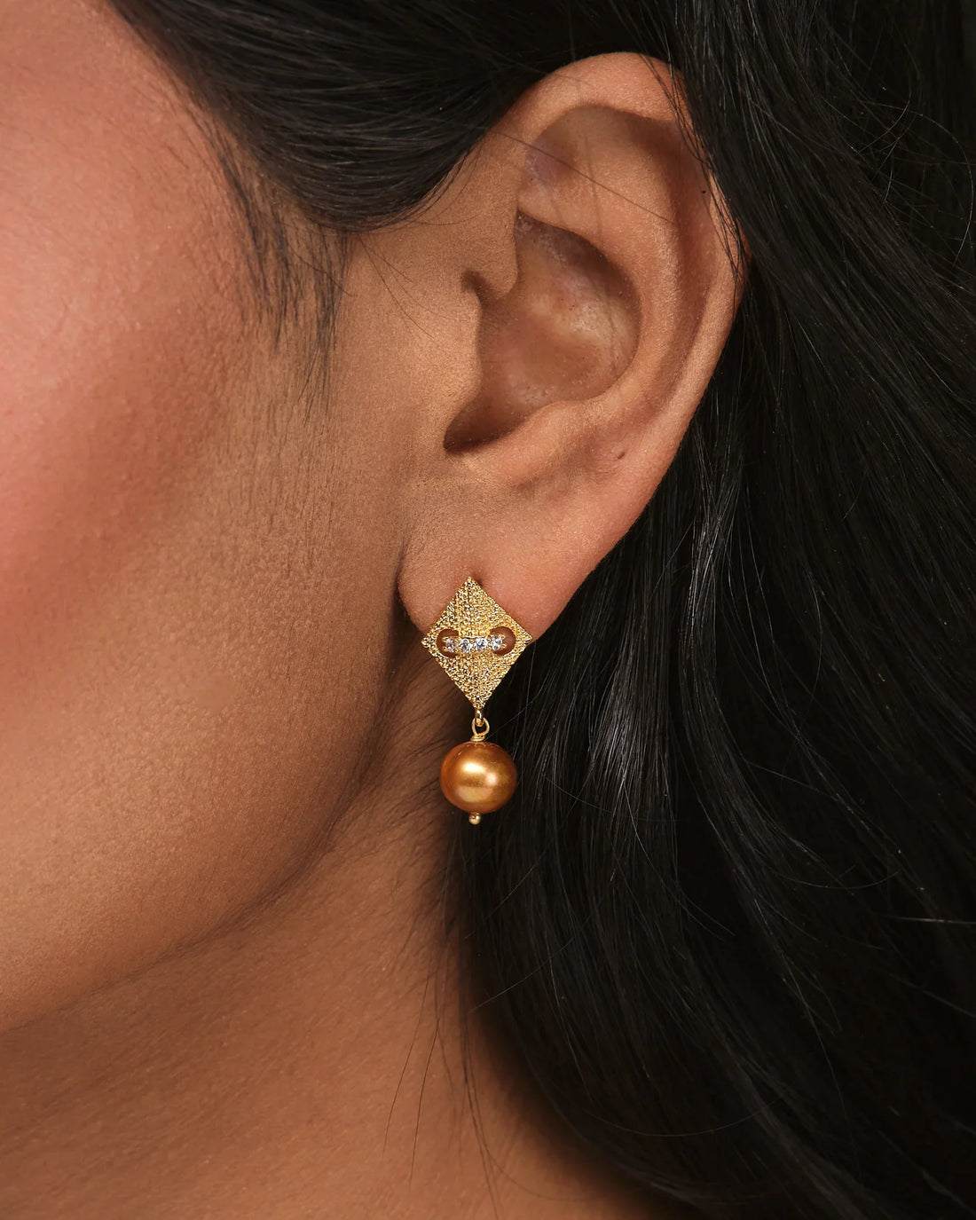 The Dainty Pearl CZ Earrings by Chandrani Pearls feature gold-toned diamond-shaped studs with clear stones and brown pearl jhumkas, shown on a beige surface with orange fabric for casual elegance.