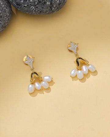 The Trendy Chandelier Jhumka with Pearls & CZ by Chandrani Pearls features clustered pearls hanging from a gold-toned, studded design, perfect for weddings or formal occasions and shown on a beige surface.