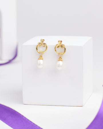 The Artsy Embrace Silver Earring by Chandrani Pearls features chic hoops with delicate bows and dangling white pearls, displayed on a white cube—perfect for elevating your style and embracing the demifine earring trend.