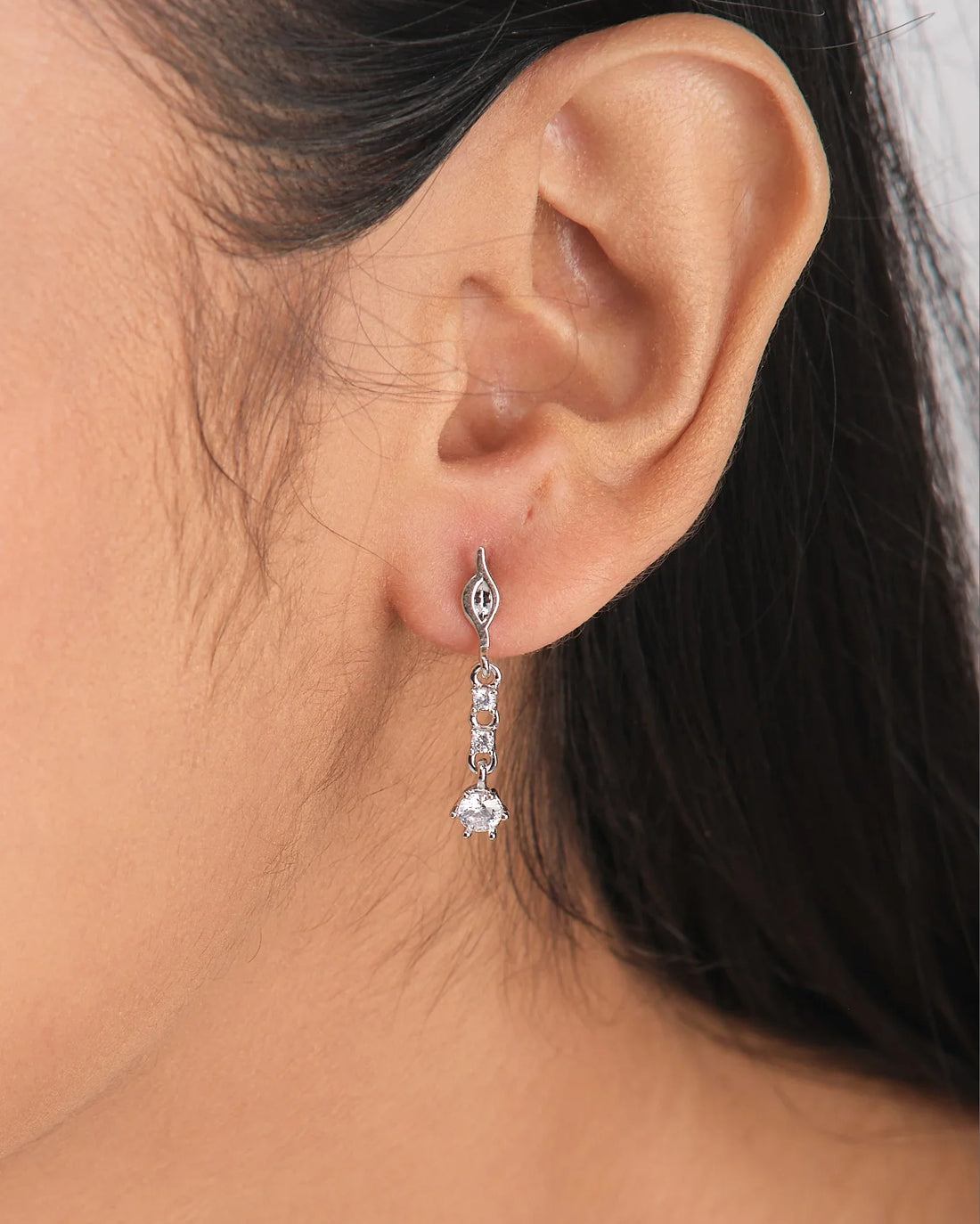 The Love Lite CZ Earrings by Chandrani Pearls are wearable silver drop earrings, each featuring three round clear CZ gemstones, shown hanging over a white bowl with a purple ribbon in the foreground.