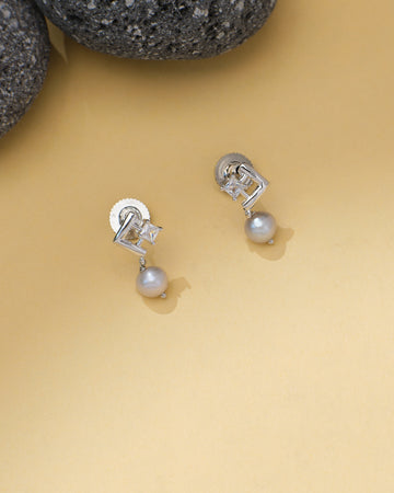 Geometric CZ and Pearl Earrings by Chandrani Pearls, featuring a silver tone, geometric design, CZ accents, and dangling pearls, displayed on a light beige surface near dark textured stones.