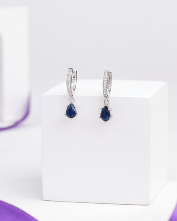 Mornings & Moonlight Silver Earrings by Chandrani Pearls feature teardrop-shaped blue gemstones and clear stones on the hoops, designed to catch the light, elegantly displayed on a white cube.