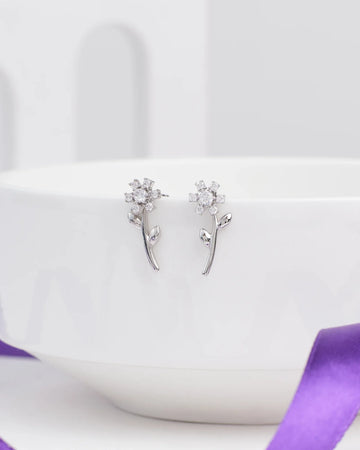 Glimmer Days Silver Studs by Chandrani Pearls—flower-shaped silver earrings with stems and leaves, ideal for daily wear—are shown resting on a white bowl, accented by a nearby purple ribbon.