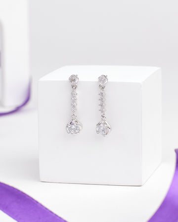 The Barely Bling Silver Earrings by Chandrani Pearls feature round and teardrop-shaped clear CZ stones for easy-care luxury, shown on a white cube with a purple ribbon beside them.
