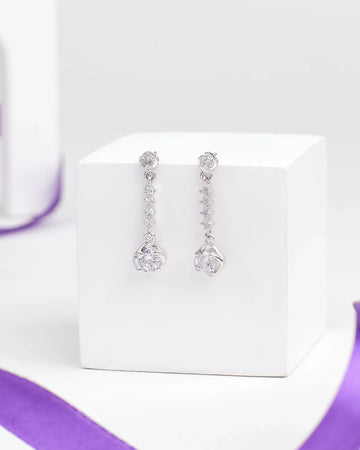 The Barely Bling Silver Earrings by Chandrani Pearls feature round and teardrop-shaped clear CZ stones for easy-care luxury, shown on a white cube with a purple ribbon beside them.