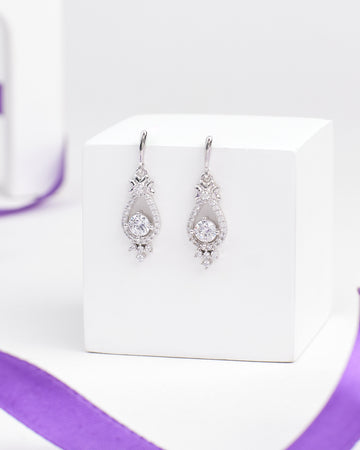 The Linger Longer CZ Silver Earrings by Chandrani Pearls feature teardrop-shaped luminous silver drops with clear gemstones, displayed on a white block and accented by a nearby purple ribbon.