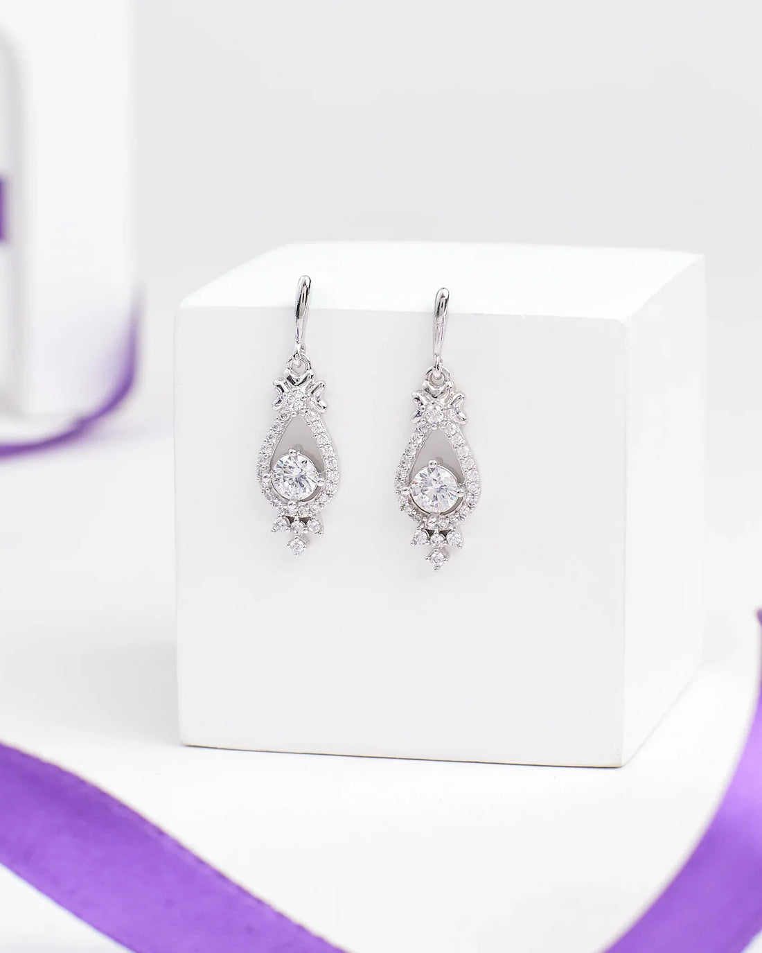 The Linger Longer CZ Silver Earrings by Chandrani Pearls feature teardrop-shaped luminous silver drops with clear gemstones, displayed on a white block and accented by a nearby purple ribbon.