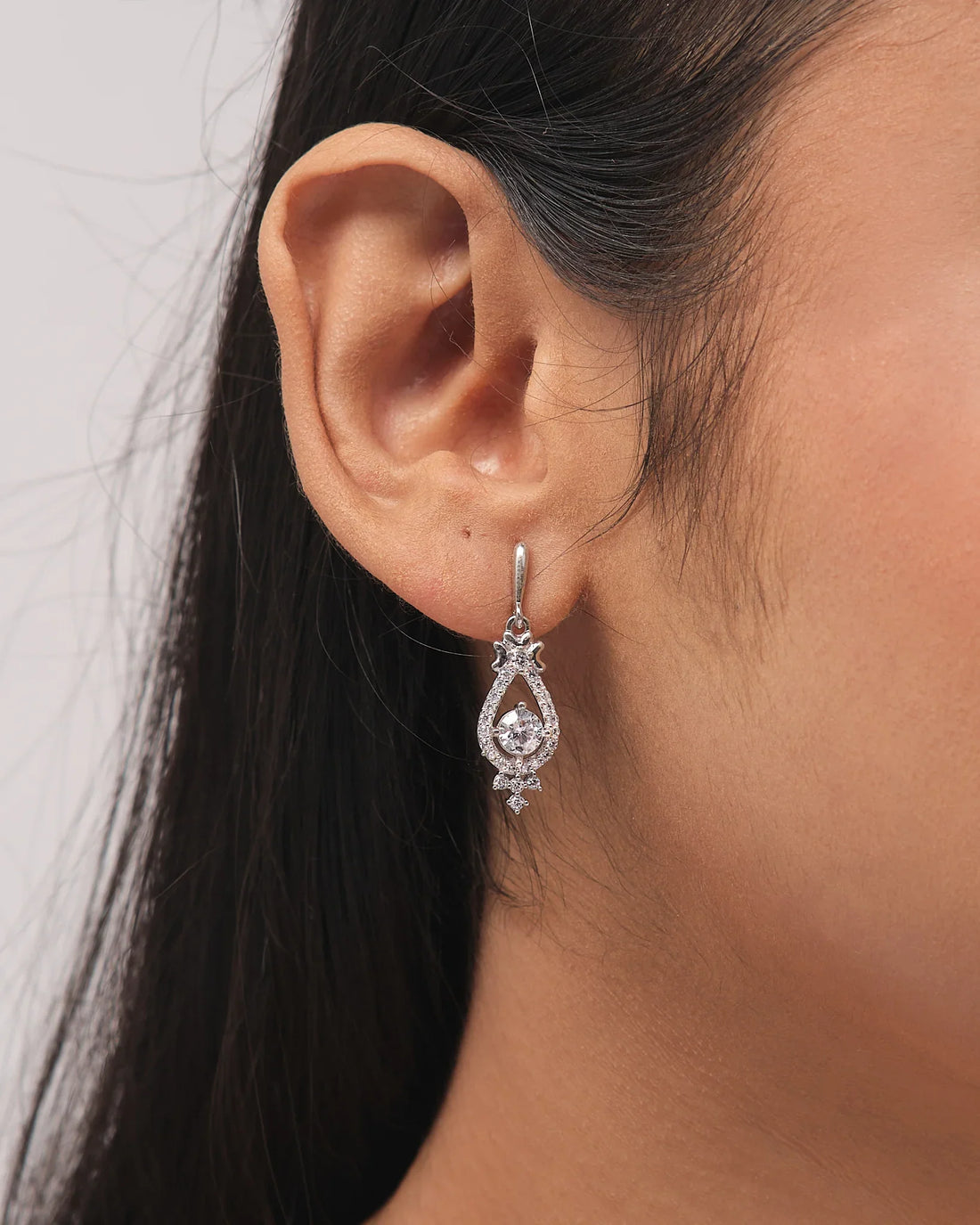 The Linger Longer CZ Silver Earrings by Chandrani Pearls feature teardrop-shaped luminous silver drops with clear gemstones, displayed on a white block and accented by a nearby purple ribbon.