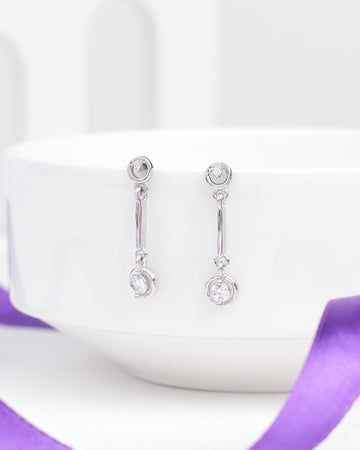The Low Key Luxe CZ Silver Earrings from Chandrani Pearls feature round clear stones and softly shine, shown on a white ceramic bowl with a purple ribbon—perfect for adding relaxed style to any look.