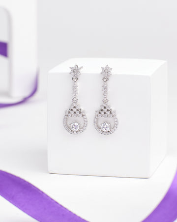 The Slow Sunday Shine Silver Earring by Chandrani Pearls features dainty silver star studs, hanging circles, and sparkling clear gemstones—an elegant accessory for any occasion. Displayed on a white box with a purple ribbon in the background.