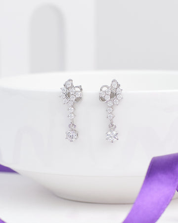 The First Kiss Glow Silver Earrings by Chandrani Pearls, featuring a romantic floral design with small clear CZ stones, are displayed hanging over the edge of a white ceramic bowl with a purple ribbon nearby.