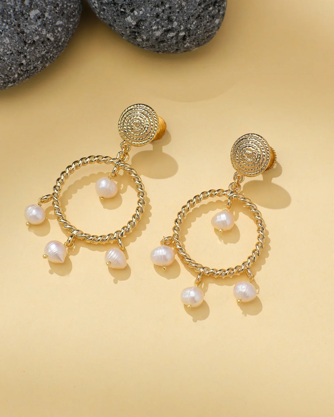 Halo Teardrop Earrings with Pearl Center by Chandrani Pearls are shown on a light yellow surface beside textured gray stones. Each gold earring features a teardrop shape with a pearl at the center.