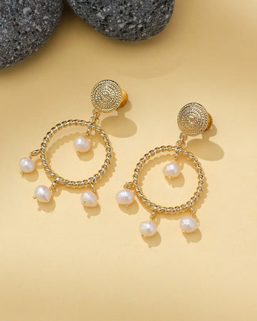 Halo Teardrop Earrings with Pearl Center by Chandrani Pearls are shown on a light yellow surface beside textured gray stones. Each gold earring features a teardrop shape with a pearl at the center.