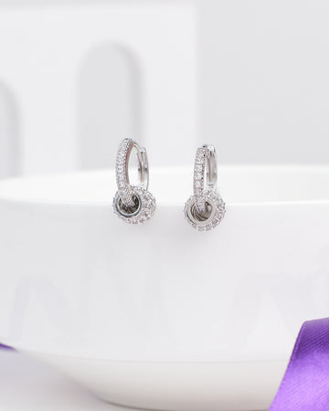 The Delicate CZ Hoop Earrings in Demifine Silver by Chandrani Pearls are lightweight, modern earrings with sparkling crystals—displayed on a white ceramic bowl and accented by a purple ribbon—ideal for any occasion.