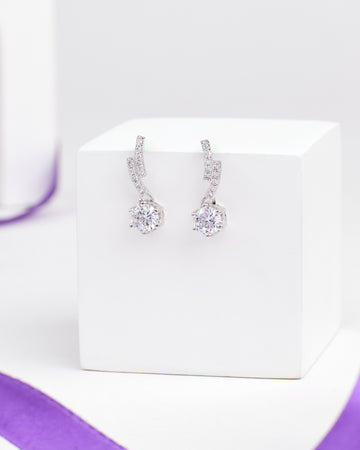 The Romantic CZ Drop Earrings in Demifine Silver by Chandrani Pearls gleam with sparkling stones, elegantly presented on a white stand with a purple ribbon backdrop.