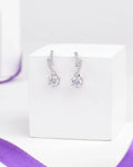 The Romantic CZ Drop Earrings in Demifine Silver by Chandrani Pearls gleam with sparkling stones, elegantly presented on a white stand with a purple ribbon backdrop.