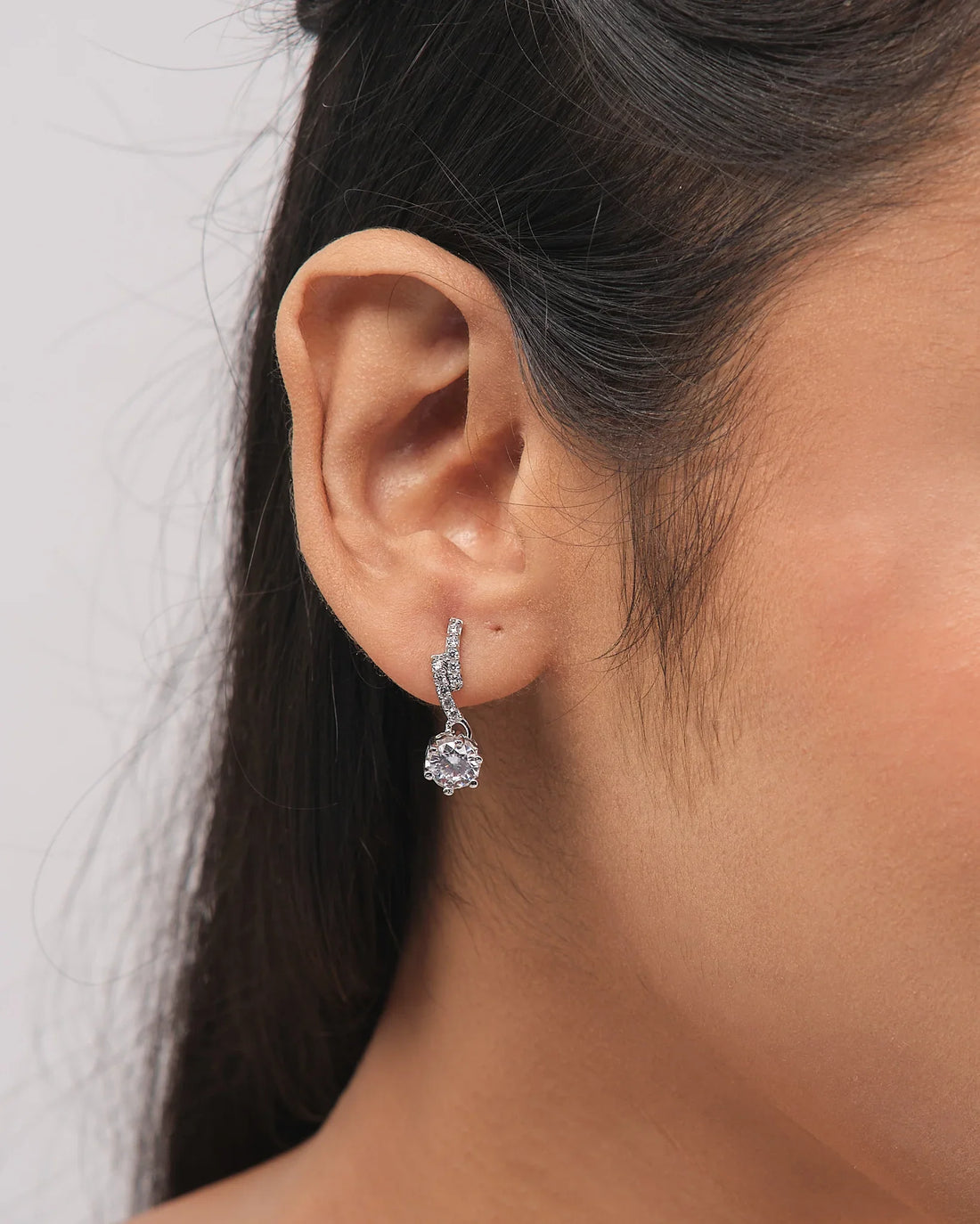The Romantic CZ Drop Earrings in Demifine Silver by Chandrani Pearls gleam with sparkling stones, elegantly presented on a white stand with a purple ribbon backdrop.