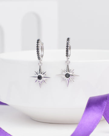 Twinkiling Inspired Demifine Silver CZ Earrings from Chandrani Pearls feature star-shaped dangles with black center stones, shown hanging from a white bowl with a purple ribbon—ideal festive jewelry.