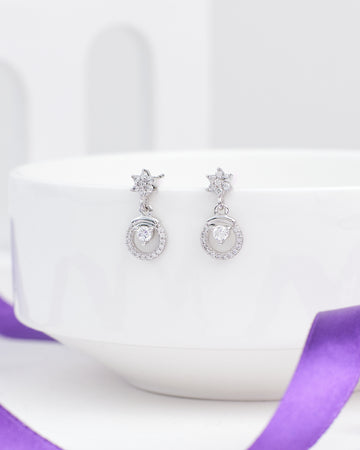 Chandrani Pearls' Floral Inspired Demifine Silver CZ Earrings, featuring star-shaped studs and gem-encrusted drops, are displayed on a white bowl with a purple ribbon—stylish festive jewelry perfect for any occasion.