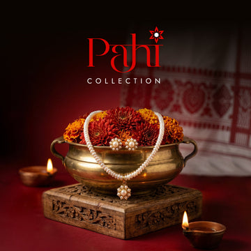 A pearl necklace and earrings set displayed on marigold flowers in a brass bowl, with two oil lamps and a patterned cloth in the background. "Pahi Collection" text above.
