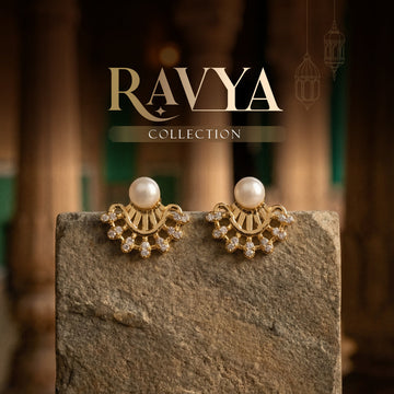 A pair of gold and pearl earrings with crystal accents displayed on a stone slab; "Ravya Collection" text appears above.