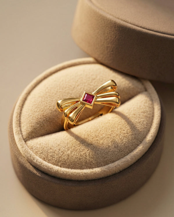 Gold bow-shaped ring with a red gemstone on a beige jewelry cushion