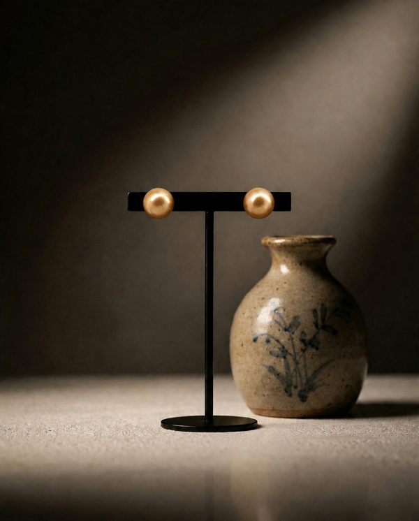 Lustrous Copper-Tone Pearl Stud Earrings by Chandrani Pearls are shown on a black stand beside a floral ceramic vase, all set on a beige surface under focused lighting.
