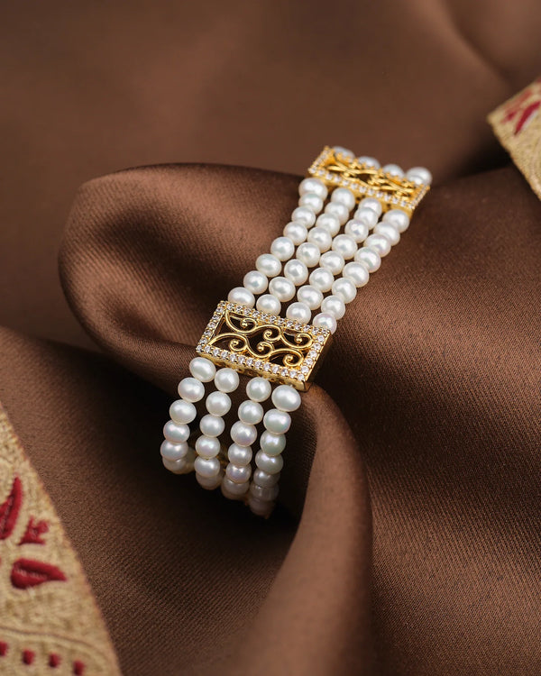 The Divine Pearl Shine Bracelet by Chandrani Pearls, a multi-strand white pearl piece with gold rectangular clasps, is elegantly displayed on brown satin fabric with embroidered trim.