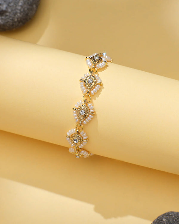 The Dainty Pearl Bracelet with CZ Halo Accents by Chandrani Pearls features teardrop-shaped charms, white pearl-like beads, and sparkling cubic zirconia, elegantly displayed on a rolled yellow surface.