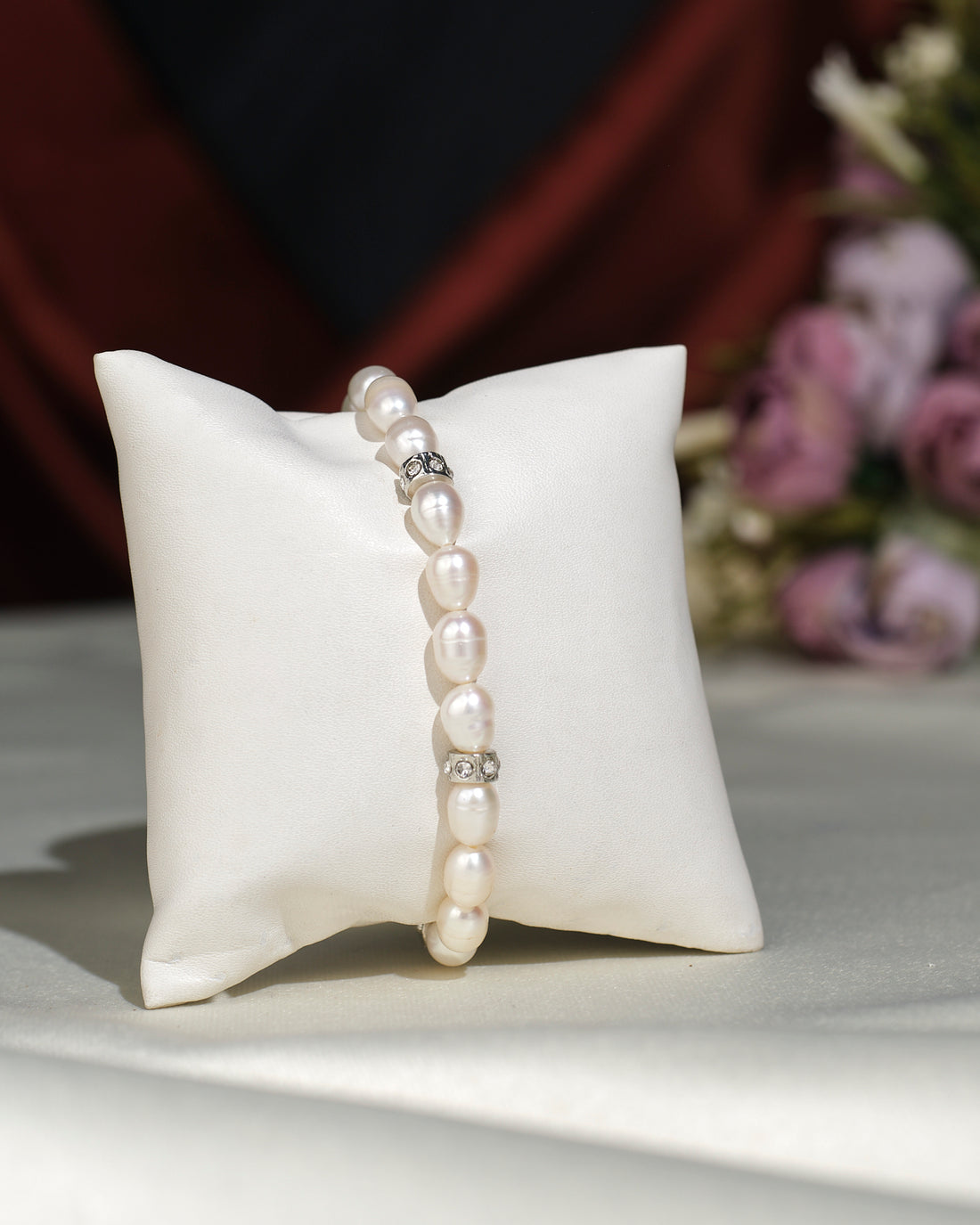 The Elegant White 1 line Bracelet by Chandrani Pearls India, symbolizing purity and elegance with silver beads, is displayed on a white pillow, with softly blurred purple flowers in the background.