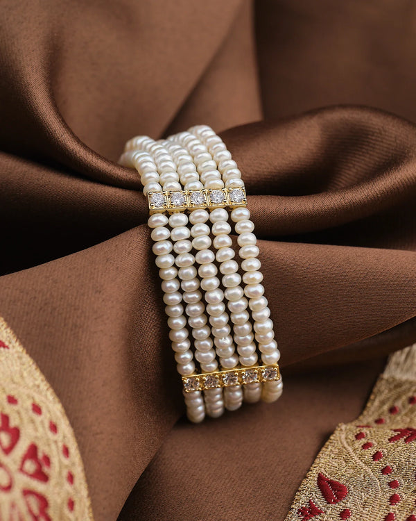 The Radiant CZ Pearl Bracelet by Chandrani Pearls features handcrafted multi-strand pearls with gold and diamond accents, beautifully displayed on brown and gold embroidered fabric.