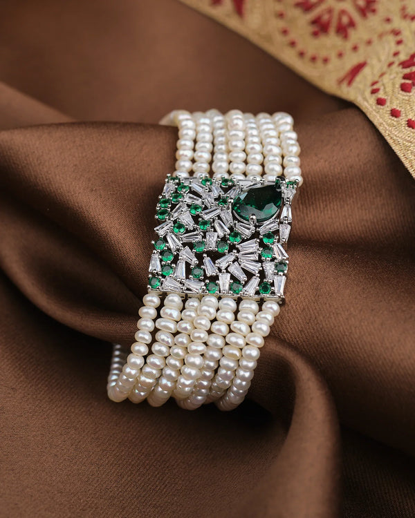 The Sparkle & Serenity Bracelet by Chandrani Pearls features multiple strands of pearls and a square silver clasp with green and clear CZ gemstones, elegantly displayed on brown fabric.