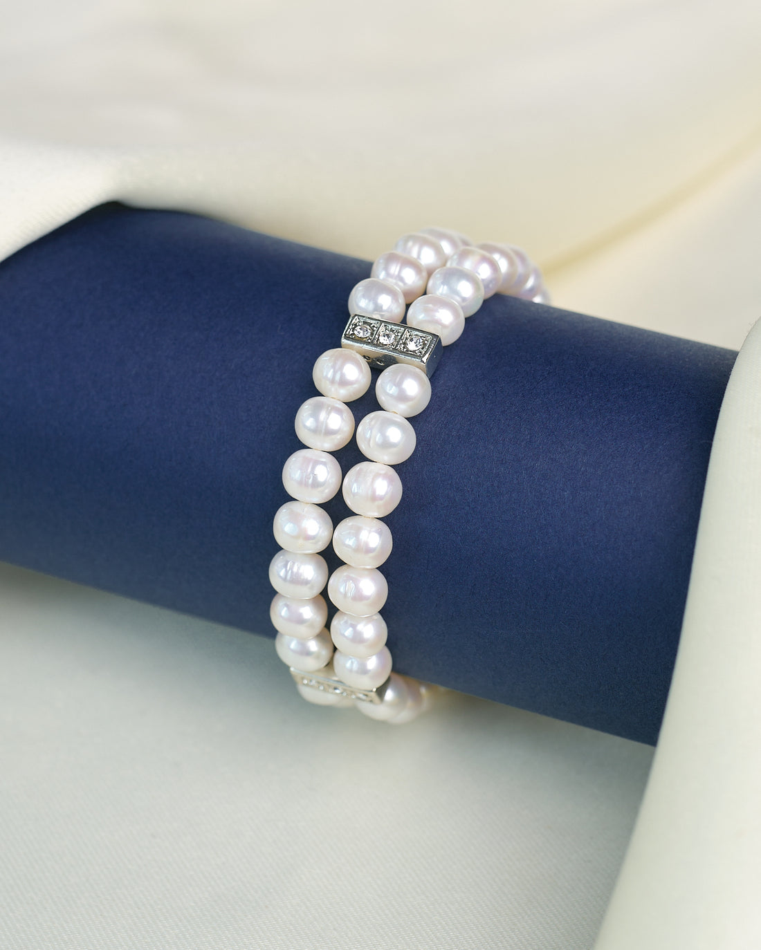 The Chandrani Pearls Elegant Pearl Bracelet with Sparkling Cubic Zirconia features double strands and rectangular silver clasps, shown on a navy stand against a cream fabric background.