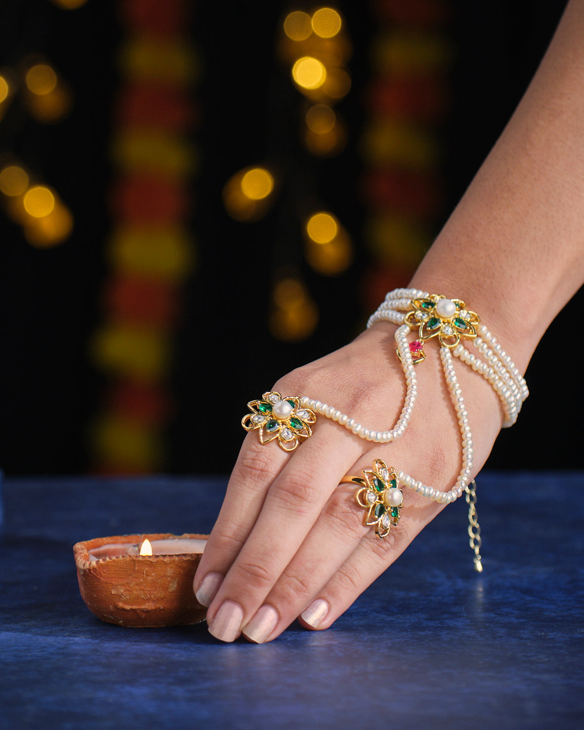 A hand wearing the Flowery Spheres Pearl Bracelet by Chandrani Pearls reaches towards a lit clay oil lamp on a blue surface, set against a softly blurred background featuring yellow and red lights.