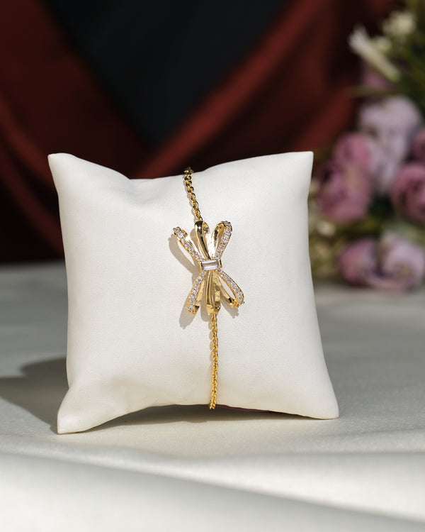 The Chandrani Pearls India Metallic CZ Pretty Bracelet, featuring a delicate bow-shaped charm adorned with CZ stones, is displayed on a white cushion with blurred flowers in the background.