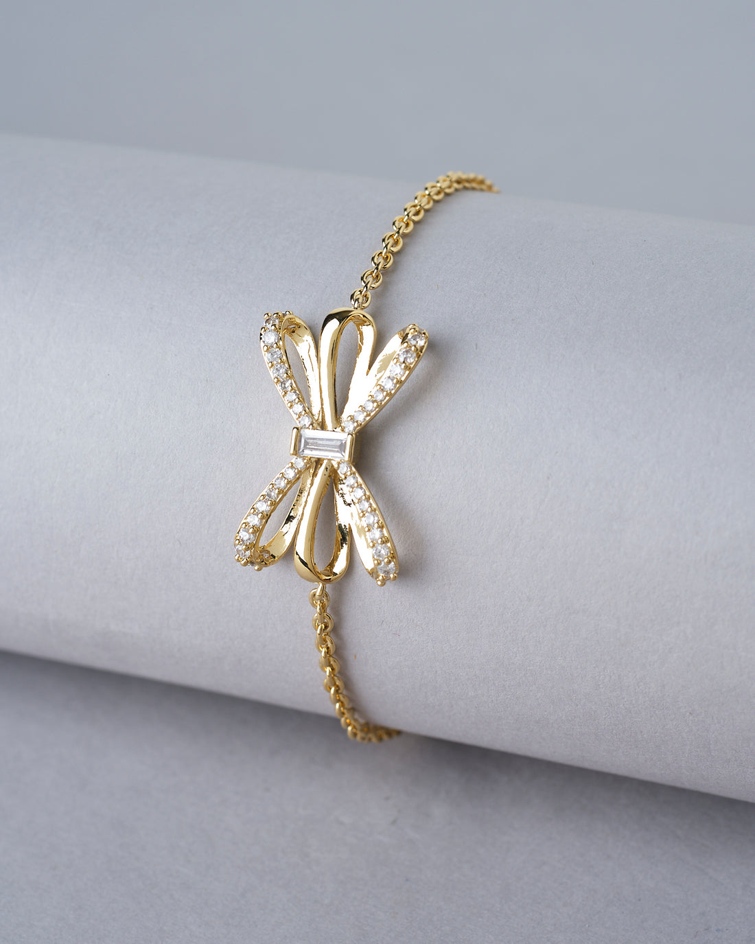 The Chandrani Pearls India Metallic CZ Pretty Bracelet, featuring a delicate bow-shaped charm adorned with CZ stones, is displayed on a white cushion with blurred flowers in the background.