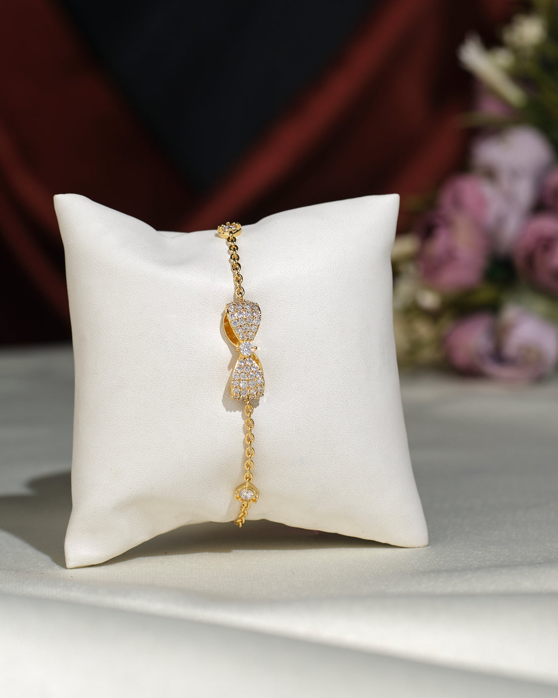 The Metallic CZ Pretty Bracelet by Chandrani Pearls India features a bow-shaped design with sparkling crystals, displayed on a white cushion with blurred flowers in the background.