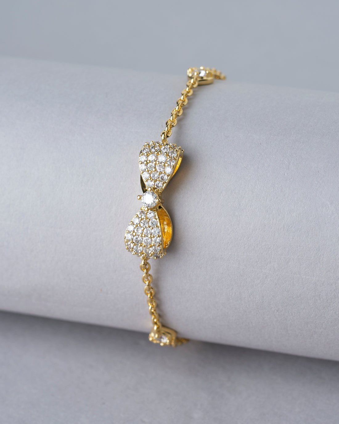 The Metallic CZ Pretty Bracelet by Chandrani Pearls India features a bow-shaped design with sparkling crystals, displayed on a white cushion with blurred flowers in the background.