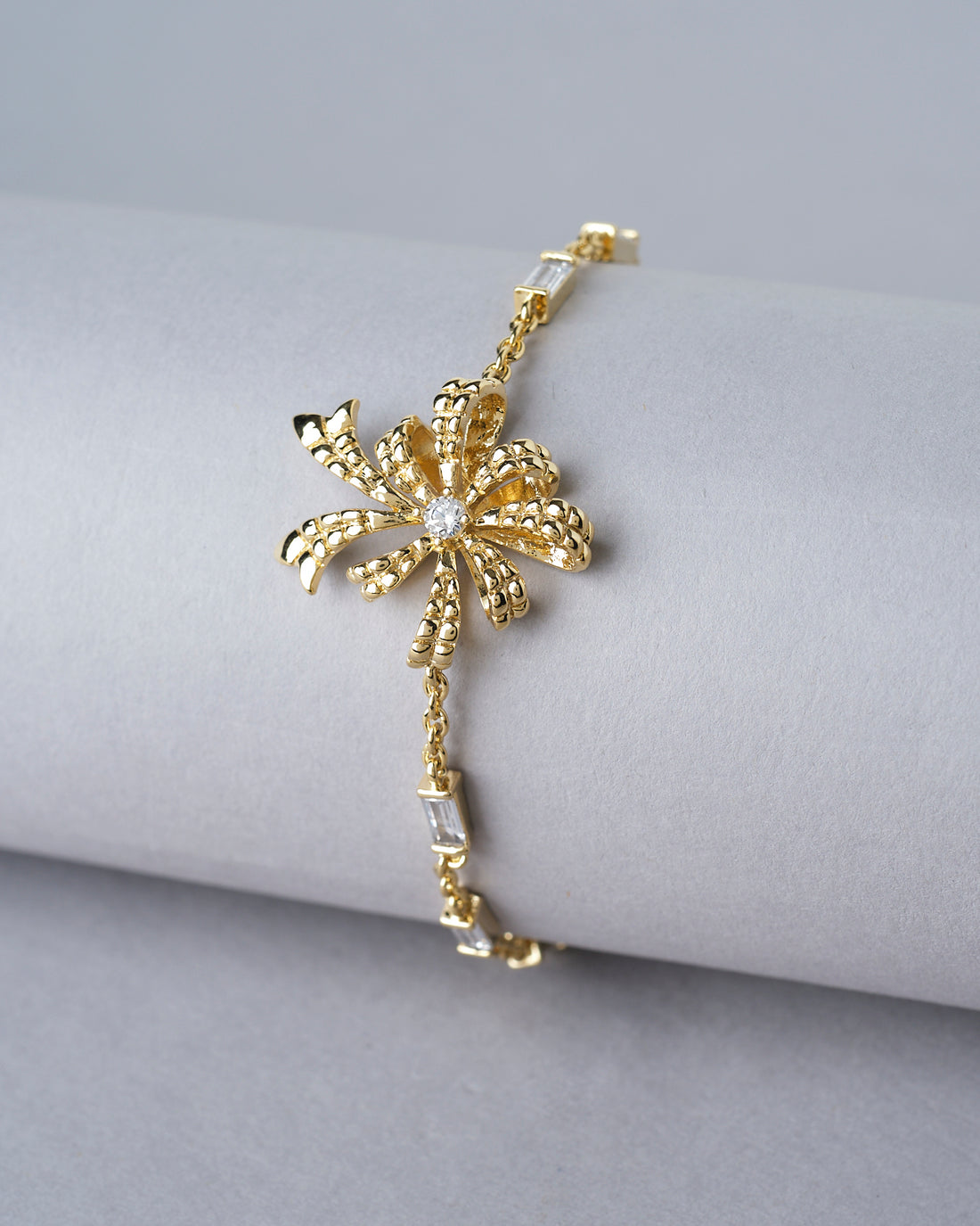 The Metallic CZ Pretty Bracelet by Chandrani Pearls India, featuring a floral design and sparkling stones, is displayed on a white cushion with blurred flowers in the background, emphasizing its unique style.