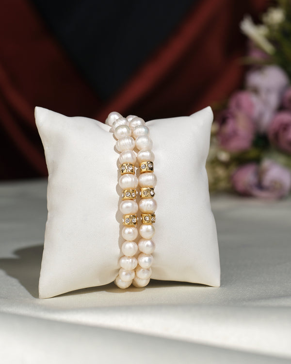 The Elegant White 2 line Bracelet by Chandrani Pearls India features double strands with gold and crystal spacers, displayed on a white cushion, with a softly blurred bouquet in the background for an elegant touch.