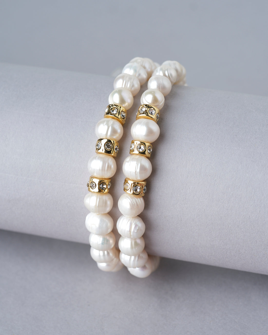 The Elegant White 2 line Bracelet by Chandrani Pearls India features double strands with gold and crystal spacers, displayed on a white cushion, with a softly blurred bouquet in the background for an elegant touch.