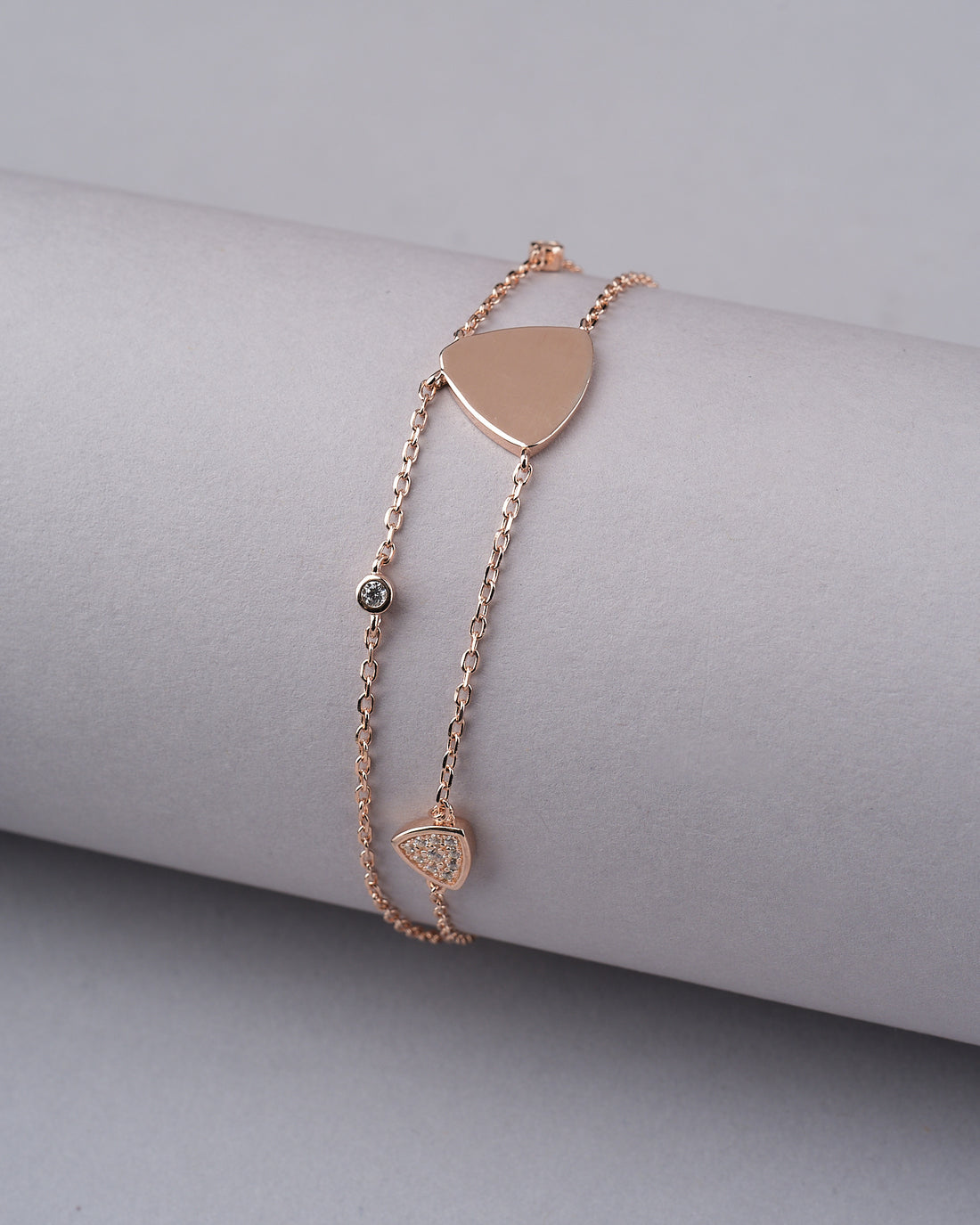 The Tamanna 925 Silver Rose Gold Bracelet from Chandrani Pearls India, featuring heart and triangle charms in a polished, unique style, is draped over a ridged cream cylinder with red roses nearby.