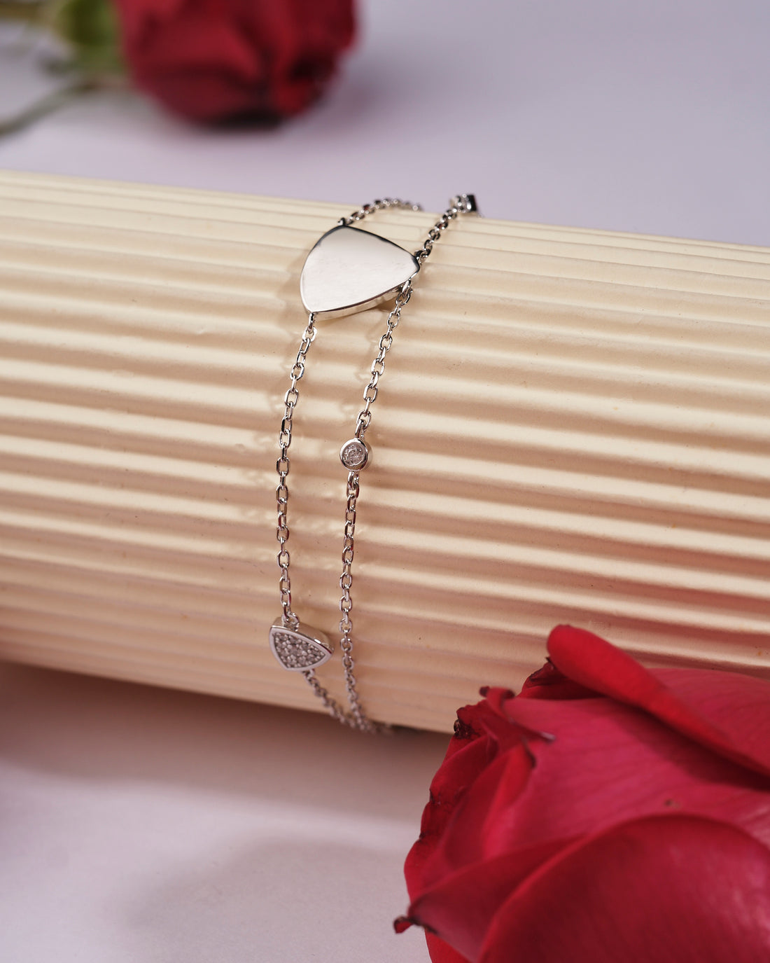 The Tvisha 925 Silver White Bracelet by Chandrani Pearls India features heart and round charms, displayed on a ribbed cylindrical surface with two red roses nearby—perfect for both casual and formal wear.