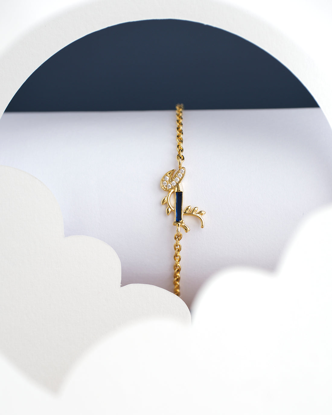 The Dainty Krishna Chain bracelet for kids by Chandrani Pearls India is displayed on a white and navy scalloped backdrop, featuring a blue gem with a leaf design.