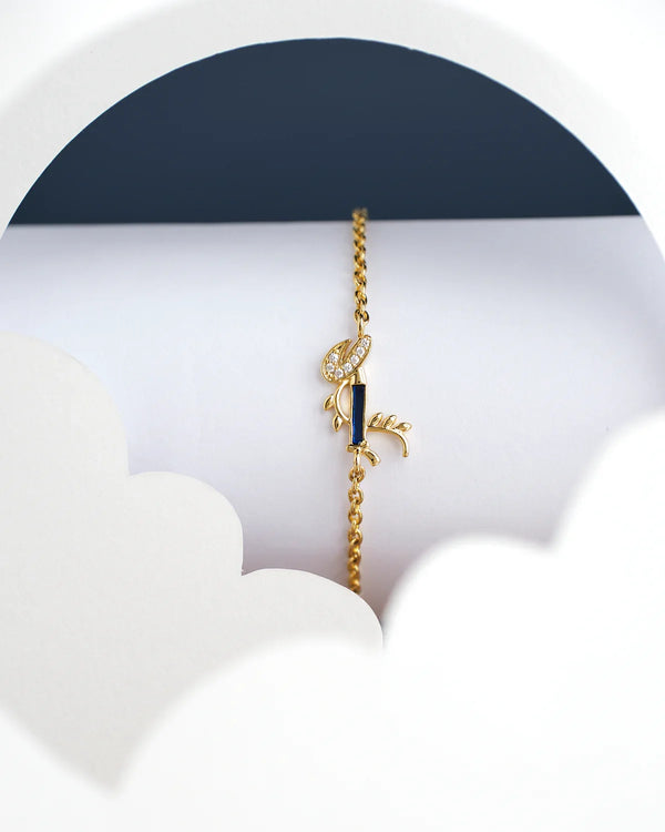 The Dainty Krishna Chain bracelet for kids by Chandrani Pearls India is displayed on a white and navy scalloped backdrop, featuring a blue gem with a leaf design.