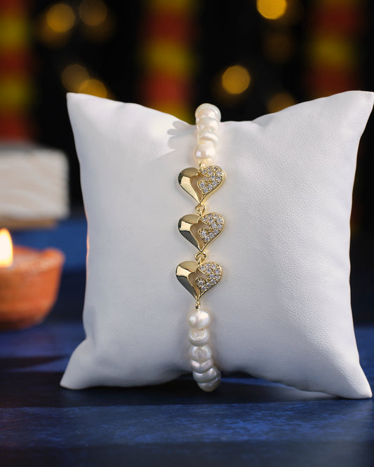 A Golden Heart Rhinestone Pearl Bracelet by Chandrani Pearls, featuring heart-shaped gold and diamond accents, is elegantly displayed on a white cushion adorned with pearl beads, set against a softly blurred background and the warm glow of a lit candle.