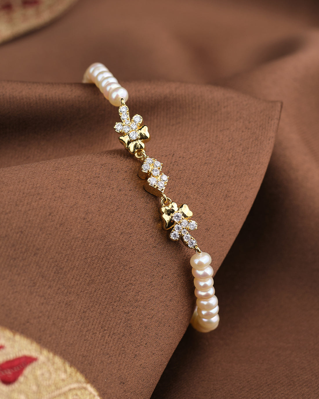 The Noor Soft Glow Bracelet by Chandrani Pearls, featuring delicate pearls and floral CZ accents, is displayed on a brown fabric background.