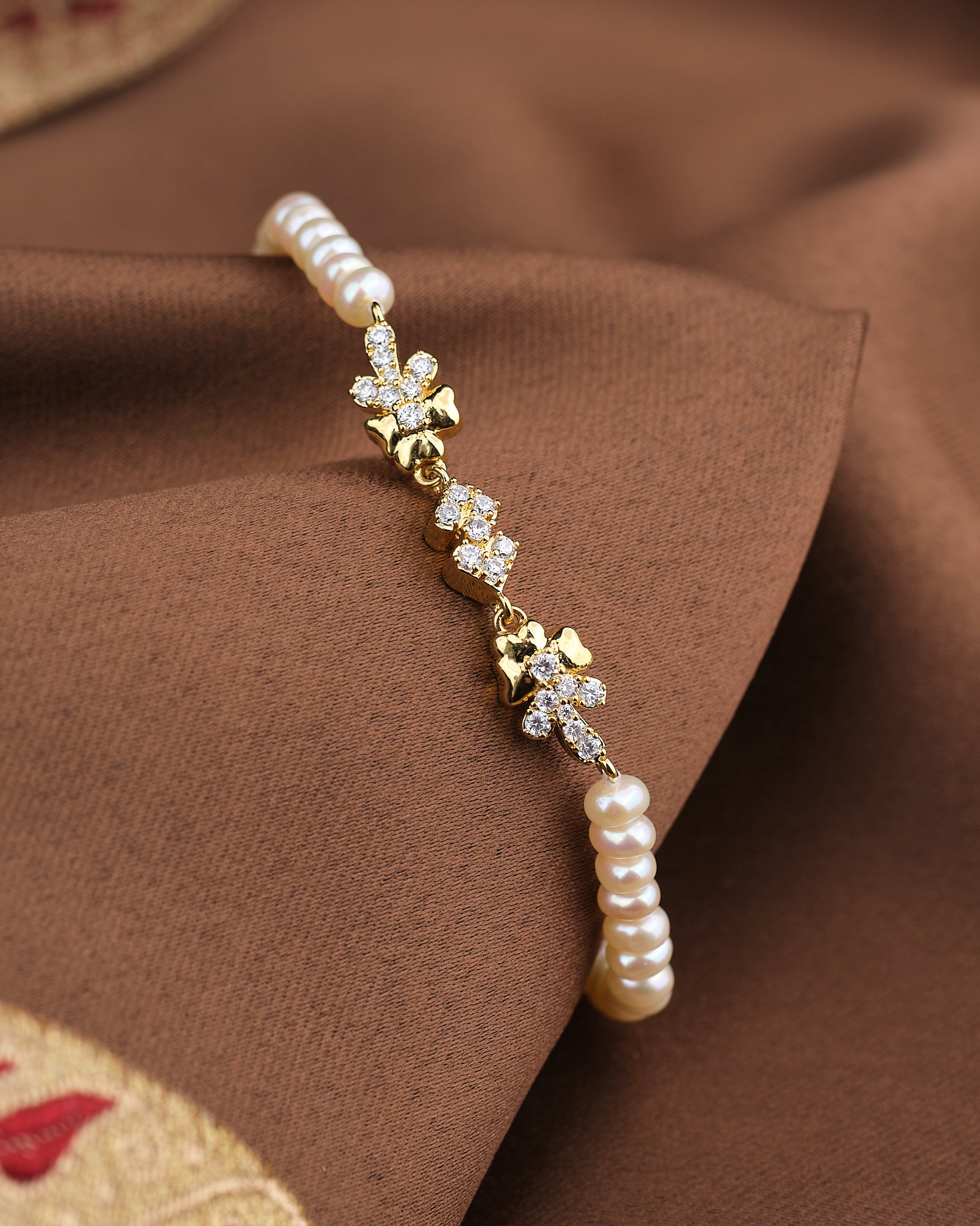 The Noor Soft Glow Bracelet by Chandrani Pearls, featuring delicate pearls and floral CZ accents, is displayed on a brown fabric background.