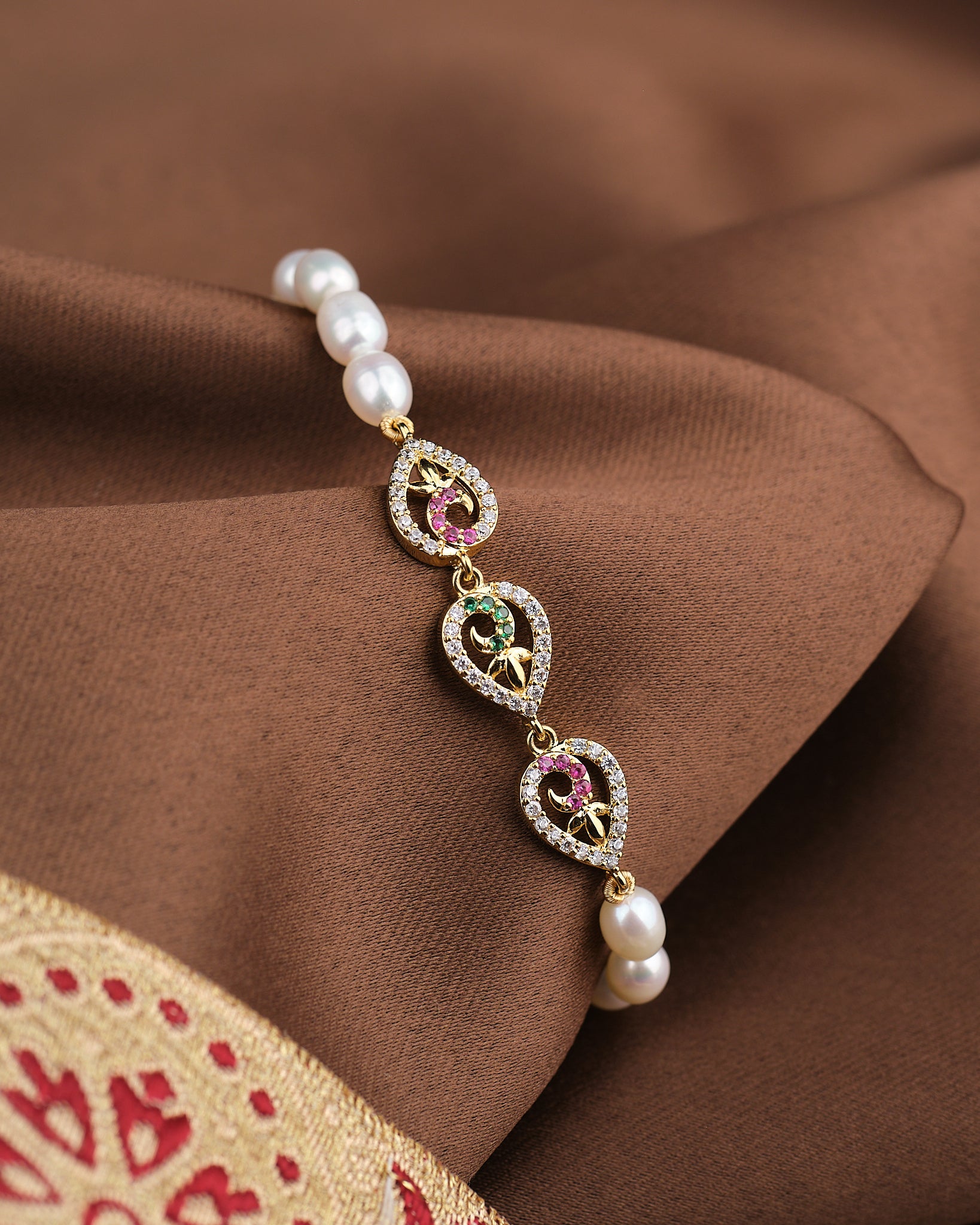 The Noor Graceful Glam Bracelet by Chandrani Pearls features heart motifs, red and green stones, and natural pearls, adding elegance to your look. Shown on brown fabric with a touch of red and gold embroidery.